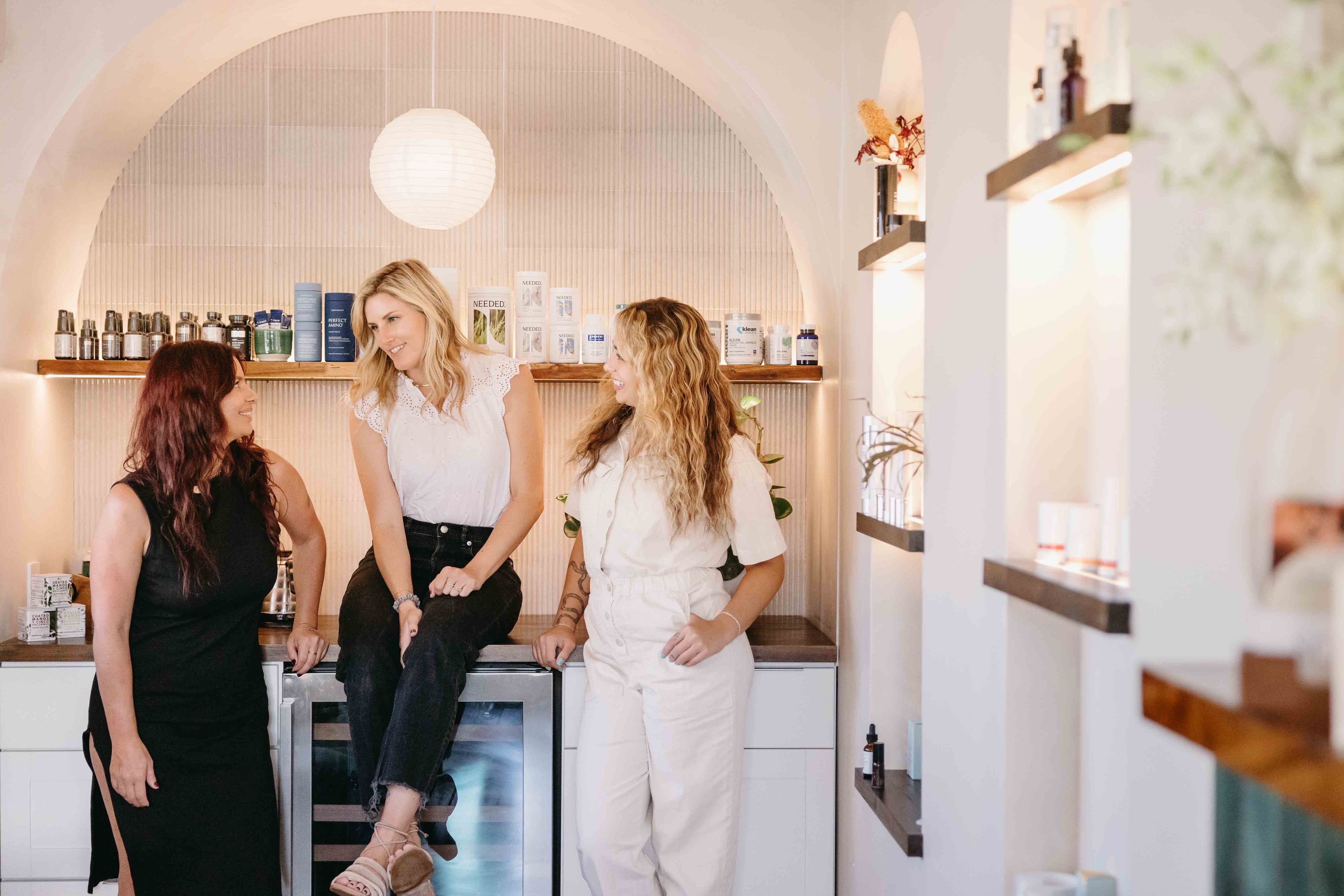 Three women sitting together in a modern interior setting with shelves and decor.