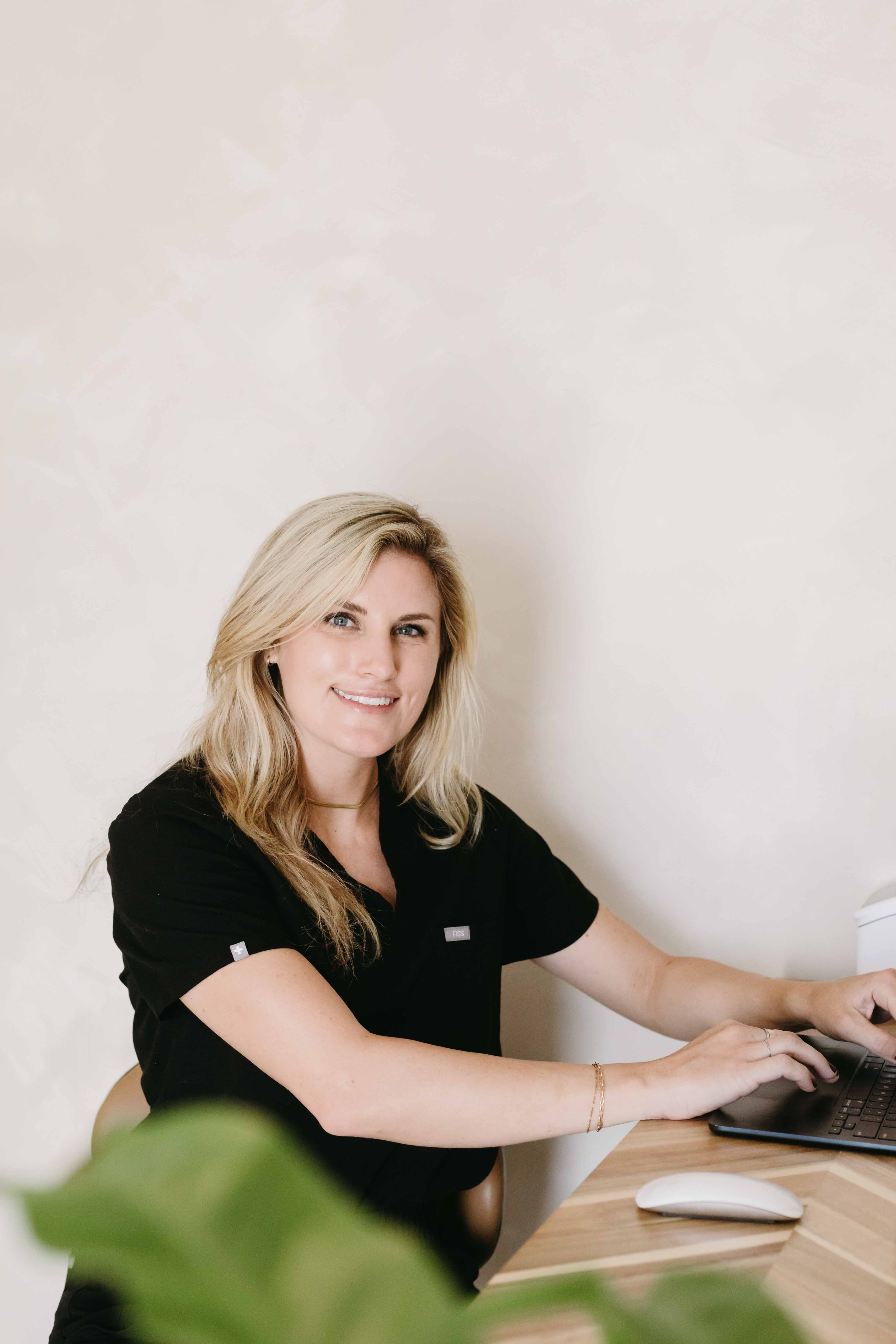 Woman in black shirt sitting at a desk with a laptop, smiling.